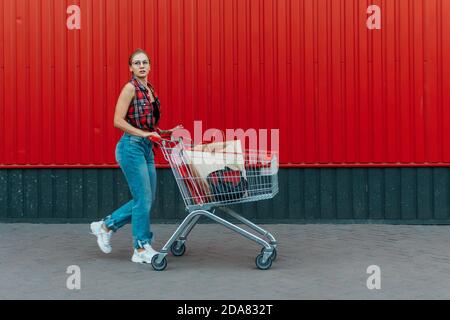 Ragazza felice con carrello di shopping su sfondo rosso del negozio di parete. Giovane donna che spinge un carrello della spesa pieno di generi alimentari e acquisti. Foto Stock