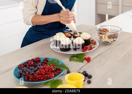 Panettiere femminile in grembiule blu decorando gustoso cupcake con cremoso piano al tavolo da cucina Foto Stock