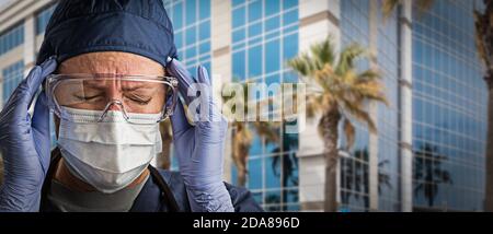 Grimacing Dottore Femminile o infermiera che indossa scrub, maschera facciale protettiva e occhiali di protezione. Foto Stock