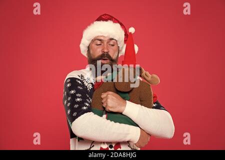 Natale concetto prep. Tanga faccia barbuta inverno maglione. Vacanze invernali. Giocattolo morbido. Abbraccio e l'amore. Acquistare regali. Inverno vendita. Festa d'inverno. La renna. L'uomo santa hat tenere regalo di Natale sfondo rosso. Foto Stock