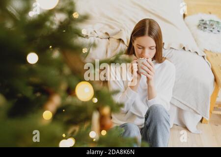 Giovane bella donna a occhi chiusi che beve cioccolata calda con marshmallow in casa decorata a natale. Foto Stock