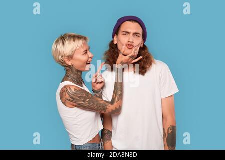 Stile libero. Giovane coppia tatuata in piedi isolata su ragazza blu rendendo grimace sul viso del ragazzo ridendo divertente Foto Stock