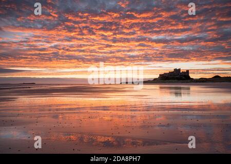 Un'alba vivida si riflette nella sabbia bagnata di Bambburgh Beach con il castello dalla silhouette adagiato su uno sperone roccioso sulla linea dell'orizzonte. Foto Stock