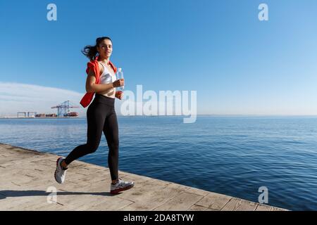 Foto all'aperto di sorridente sportivo che fa jogging e si allenano sul lungomare. Fitness donna che corre e guardando la macchina fotografica Foto Stock