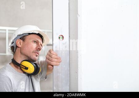 uomo lavoratore guarda il livello di spirito controlla la parete con elmetto e cuffie di protezione dell'orecchio, parete bianca con spazio di copia Foto Stock