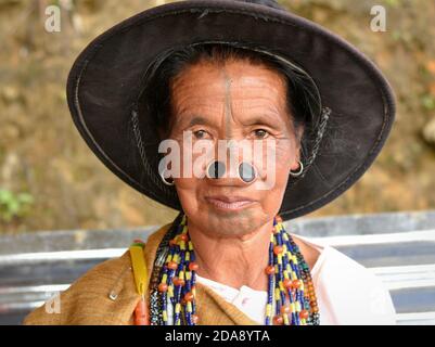 Anziana tribale dell'India nordorientale Apatani con tappi neri al naso e tatuaggi tradizionali del viso indossa un cappello moderno e posa per la macchina fotografica. Foto Stock
