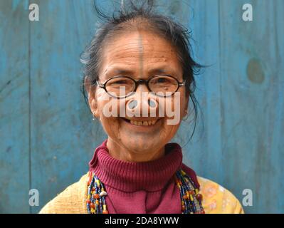 La vecchia tribale dell'India nordorientale Apatani con i tappi neri del naso di legno e il tatuaggio tradizionale del viso indossa gli occhiali moderni e sorrisi per la macchina fotografica. Foto Stock