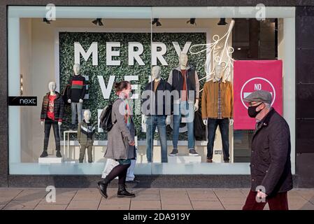 Gli acquirenti che indossano maschere facciali camminano vicino alla vetrina del negozio Next su Princes Street, Edimburgo, Scozia, Regno Unito. Foto Stock