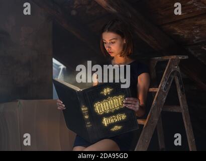 Ragazza attraente in abito nero che legge libro mistico in soffitta Foto Stock