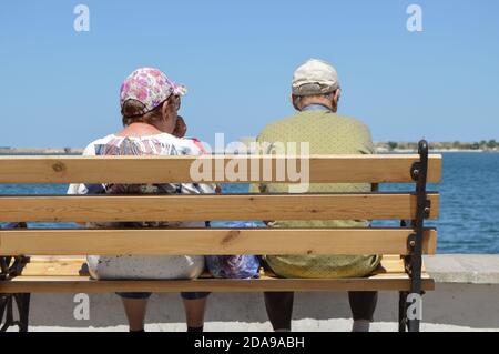 Una coppia anziana è seduta su una panchina vicino al mare e ammirando il mare, la vista dal retro Foto Stock