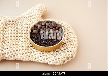 vasetto con scrub al caffè e grani su guanto con spazzola per massaggio ruvido in iuta su fondo beige pulito, layout cosmetico per la cura del corpo a casa, fuoco selettivo Foto Stock