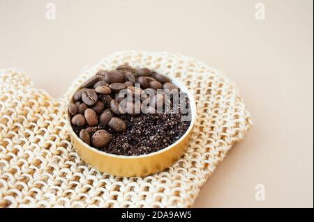 vasetto con scrub al caffè e grani su guanto con spazzola per massaggio ruvido in iuta su fondo beige pulito, layout cosmetico per la cura del corpo a casa, fuoco selettivo Foto Stock