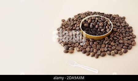 recipiente rotondo con caffè tritato per lo scrub e spatola per l'applicazione circondata da chicchi di caffè su sfondo beige. Layout cosmetico di cura del corpo domestico, se Foto Stock