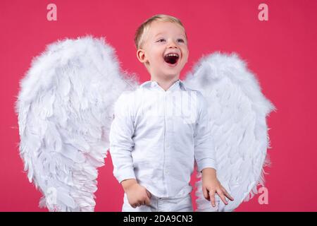 Ritratto di felice sorridente piccolo biondo riccamente Angel boy. Bambino angelico. Bambino che indossa angelo vestito bianco costume e ali di piume. Consept di Foto Stock