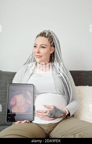 Ritratto di felice giovane donna sorridente con i Dreadlock seduti sopra divano e mostra l'immagine ecografica del bambino Foto Stock
