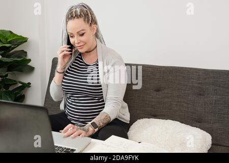 Bella giovane donna incinta che parla al telefono con la collega quando lavorare su un computer portatile a casa Foto Stock