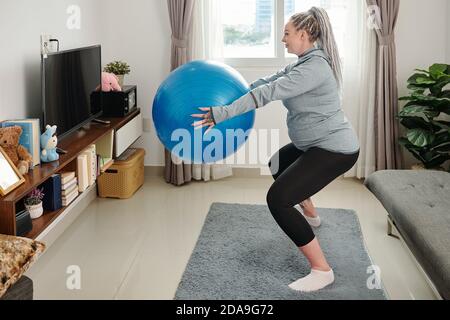 Donna giovane incinta sorridente che fa squats con palla di idoneità dentro le mani Foto Stock