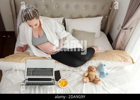 Donna incinta che riposa sul letto, mangia frutta tagliata e guarda film sullo schermo del computer portatile Foto Stock