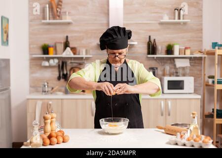 Donna anziana che prepara l'impasto cracking uova su farina di grano seguendo la ricetta tradizionale. Cuoco di pasta anziano che coglie l'uovo sulla ciotola di vetro per la ricetta della torta in cucina, mescolando a mano, impastando. Foto Stock