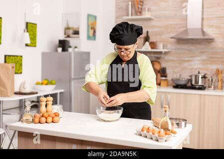 La donna rompe l'uovo sopra la farina facendo l'impasto per i prodotti da forno. Anziano pasticciere spezzare l'uovo su una ciotola di vetro per la ricetta della torta in cucina, mescolando a mano, impastando gli ingredienti prreparing torta fatta in casa Foto Stock