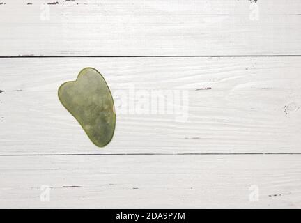 Raschietto di gouache fatto di pietra verde naturale su uno sfondo di legno bianco. Massaggio anti-invecchiamento, cura della pelle. Spazio libero per il testo, vista dall'alto. Foto Stock