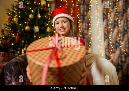 Un bambino felice nel cappello rosso di Santa sta celebrando il Natale. Carina ragazza che tiene avvolto dono, sorridente e ondeggiante mano, seduta in decorato con Natale Foto Stock