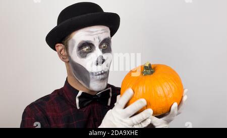 Un pagliaccio spaventoso guarda la macchina fotografica, tiene una zucca nelle mani e la minaccia con un coltello. Costume di Halloween. Foto Stock