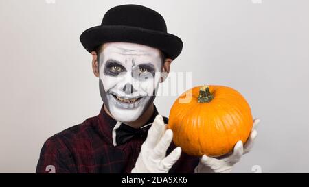 Un pagliaccio spaventoso guarda la macchina fotografica, tiene una zucca nelle mani e la minaccia con un coltello. Costume di Halloween. Foto Stock