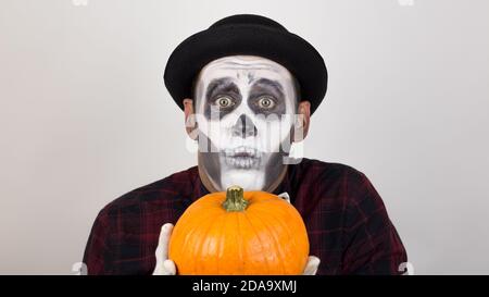 Un pagliaccio spaventoso guarda la macchina fotografica, tiene una zucca nelle mani e la minaccia con un coltello. Costume di Halloween. Foto Stock