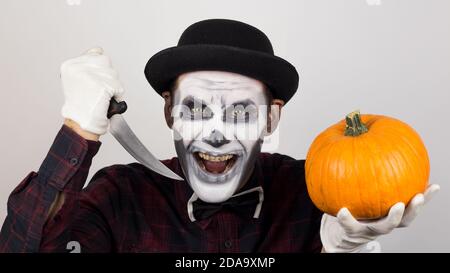 Un pagliaccio spaventoso guarda la macchina fotografica, tiene una zucca nelle mani e la minaccia con un coltello. Costume di Halloween. Foto Stock