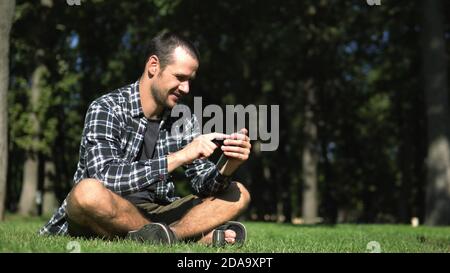 Giovane bel ragazzo in una camicia plaid foglie attraverso il feed di social network mentre si siede sull'erba nel parco. Un ragazzo giovane legge il feed di Foto Stock