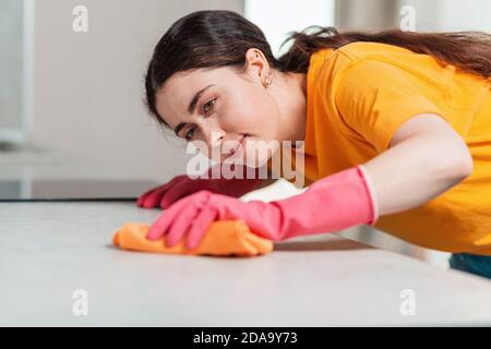 Una giovane donna in guanti di gomma strofina il tavolo con uno straccio. Primo piano verticale dal lato. Pulizia della casa. Foto Stock