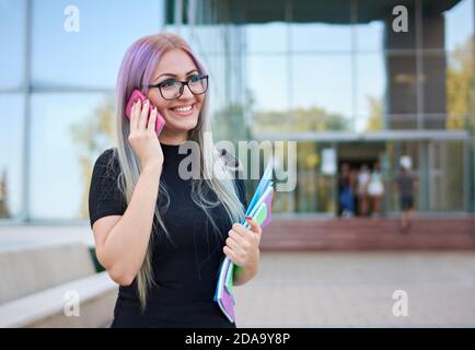 Sorridente giovane studentessa di generazione z in piedi in un campus di fronte alla facciata di un edificio universitario e che parla sullo smartphone Foto Stock