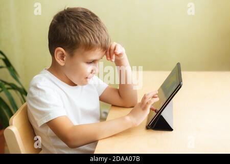 Carino scolaro che fa i compiti con il computer a casa. Figlio che utilizza i gadget per lo studio. Concetto di istruzione e formazione a distanza. Homeschooling durante la quarantena. Intrattenimento domestico. Foto Stock