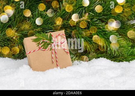 Regalo di Natale senza plastica fatto a mano sotto l'abete. Confezione regalo avvolta in carta artigianale e arco di corda si erge nella neve con rami di pino e bokeh Foto Stock