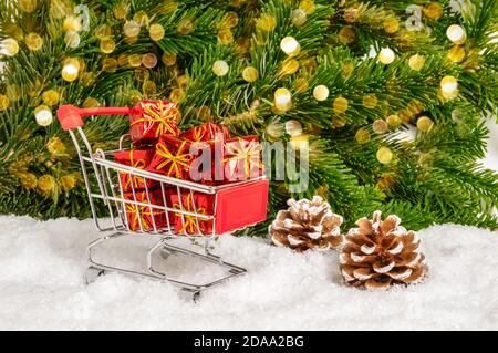 Shopping per regali di Natale. Closeup di carrello rosso e molti piccoli regali in neve con coni di pino, abete e bokeh luci sfondo. Wint Foto Stock