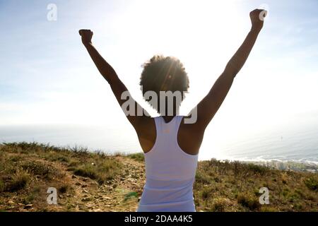 Vista posteriore ritratto di giovane donna in piedi su scogliera con le mani alzate verso il cielo e guardando il mare Foto Stock