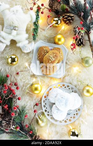 Melomakarona e kourabiedes, dolci tradizionali greci di Natale, luci di natale, palle, una bambola di babbo natale e rami artificiali di pino su pelle di pecora Foto Stock