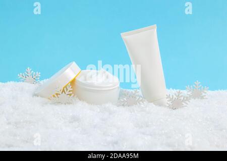 Aprire il vaso crema viso e crema mani o tubo lozione corpo in neve e fiocchi di neve su sfondo blu. Prodotti cosmetici invernali per la cura della pelle Foto Stock