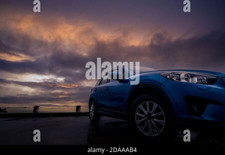 Auto SUV di lusso con gocce di pioggia. Vista frontale nuova auto blu SUV parcheggiata su un parcheggio in cemento vicino alla spiaggia con cielo al tramonto. Concetto di veicolo elettrico. RO Foto Stock