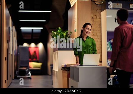 Giovane allegra donna asiatica che parla con la collega mentre si sta insieme nel corridoio dell'ufficio moderno. Uomini d'affari e concetto di lavoro Foto Stock