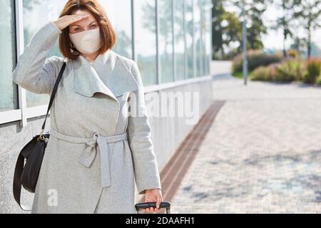 Donna d'affari con valigia e maschera a causa della pandemia di Covid-19 in viaggio d'affari Foto Stock