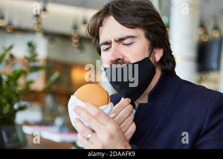 Uomo con una maschera facciale sul mento a causa di Covid-19 mangiare un hamburger nel ristorante Foto Stock