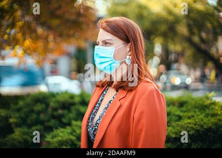 Coronavirus, covid 19 concetto. Giovane ragazza in una maschera medica e tuta tiene uno smartphone nelle mani e guarda a lato sulla strada Foto Stock