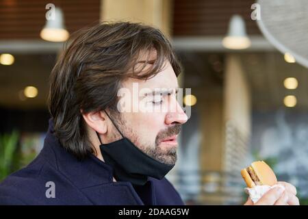 Uomo con una maschera sul mento a causa di Covid-19 mentre si mangia un hamburger nel ristorante snack Foto Stock
