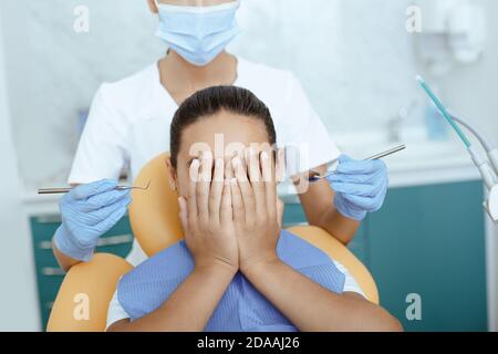 Spaventato ragazza latina in sedia dentale copre faccia e. non dà la giovane donna medico esame denti in clinica Foto Stock