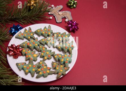 Piatto bianco con pan di zenzero di Natale sotto forma di alberi di Natale, rami di pino, giocattoli di Natale su uno sfondo rosso.primo piano, spazio di copia Foto Stock