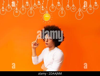 Ragazzo nero intelligente che punta il dito verso l'alto sulle lampadine su sfondo arancione, collage Foto Stock