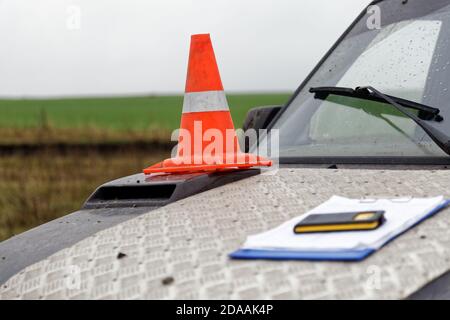 Cono stradale arancione con striscia bianca sul cofano di un veicolo per il servizio stradale. Clip per supporto carta per Appunti e telefono in primo piano sfocato. Poco profondo Foto Stock