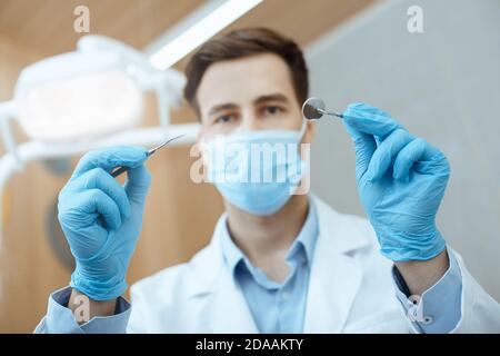 Stomatologo millenario amichevole in camice bianco, maschera protettiva e guanti di gomma che lavora con attrezzature professionali Foto Stock